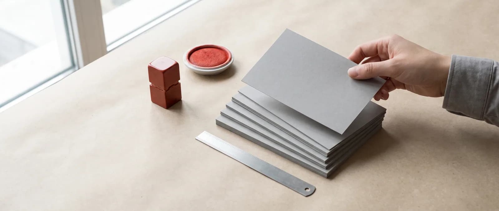 A hand lifting the top card off a stack of blank grey cards, beside an upright red seal standing on its carved face and an ink pad.