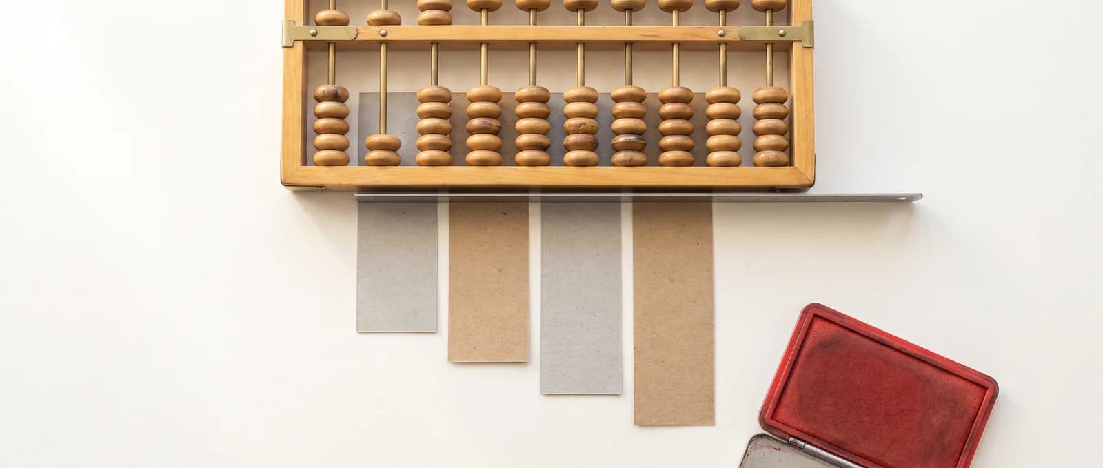 A pale wooden abacus lying above four blank paper slips arranged in a descending stagger, held straight by a steel rule.