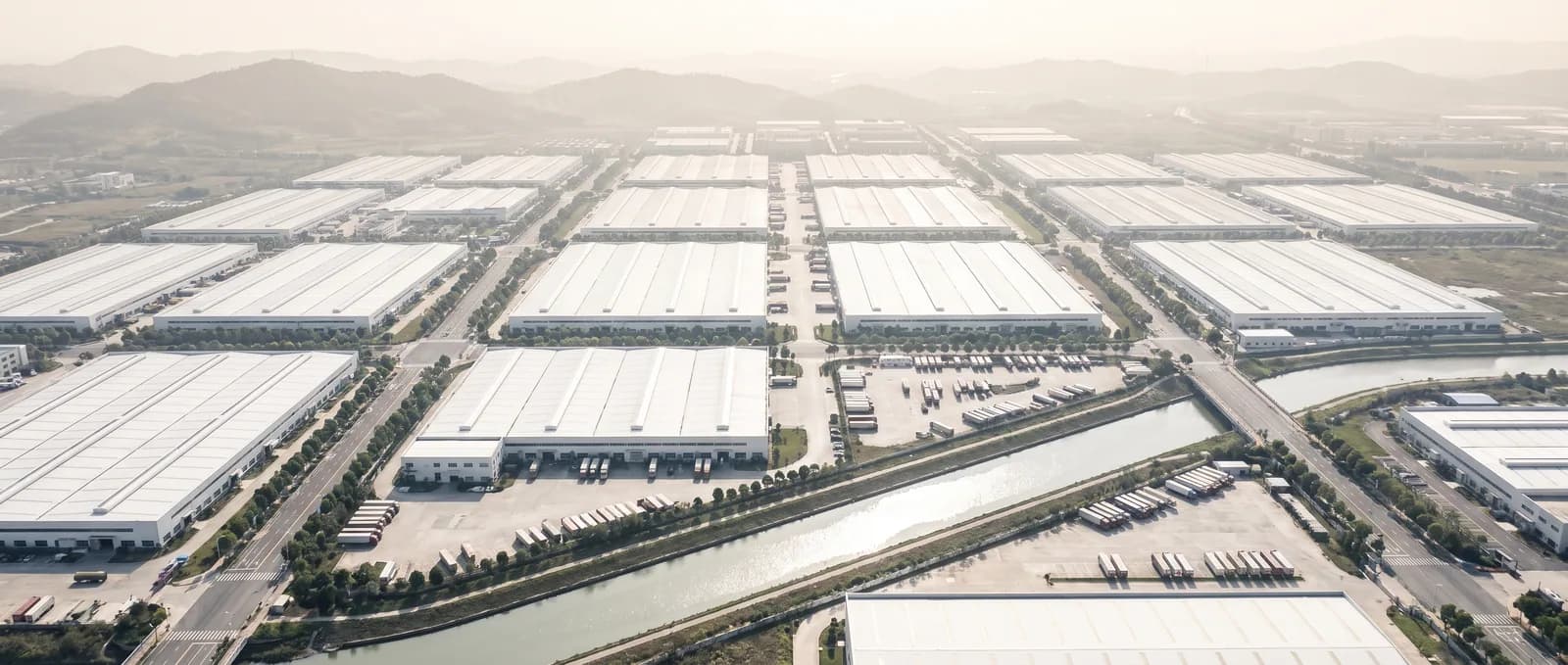 An aerial view over a grid of white-roofed factory halls, access roads and loading yards beside a river channel.